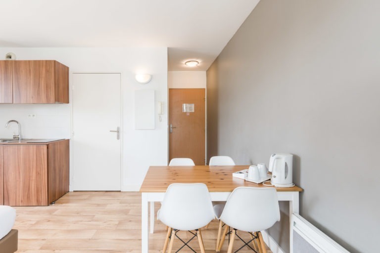 L'espace repas moderne et minimaliste des Appartements De Vacances Bordeaux comprend une table en bois avec quatre chaises blanches. La table contient une bouilloire et un plateau. À gauche, se trouve une kitchenette compacte avec des armoires en bois et un évier. L'espace est doté d'un parquet en bois clair et de murs de couleur neutre avec une porte blanche menant à une autre pièce.