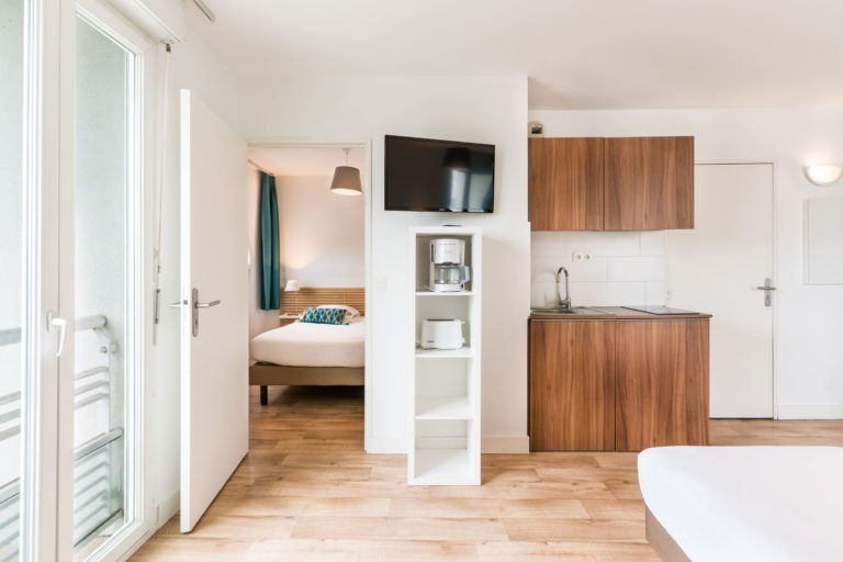 Appartement moderne avec une chambre confortable avec un grand lit et des rideaux turquoise. À côté, un espace ouvert comprend une cuisine compacte avec des armoires en bois, des étagères blanches contenant une cafetière et un grille-pain, et une télévision montée au-dessus. Une porte-fenêtre est visible sur la gauche, typique des Appartements De Vacances Bordeaux.