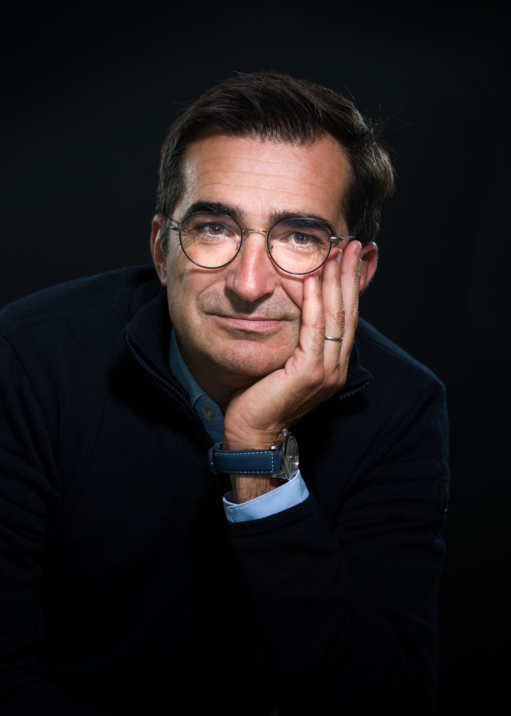 Portrait d'un homme d'âge moyen aux cheveux courts et foncés, portant des lunettes rondes, une veste zippée foncée et une chemise bleue. Il pose sa tête sur sa main gauche, montrant une bague. Son expression est contemplative, avec un léger sourire. L'arrière-plan est sombre, mettant en valeur son visage et ses lunettes, parfait pour les portraits professionnels à Bordeaux.