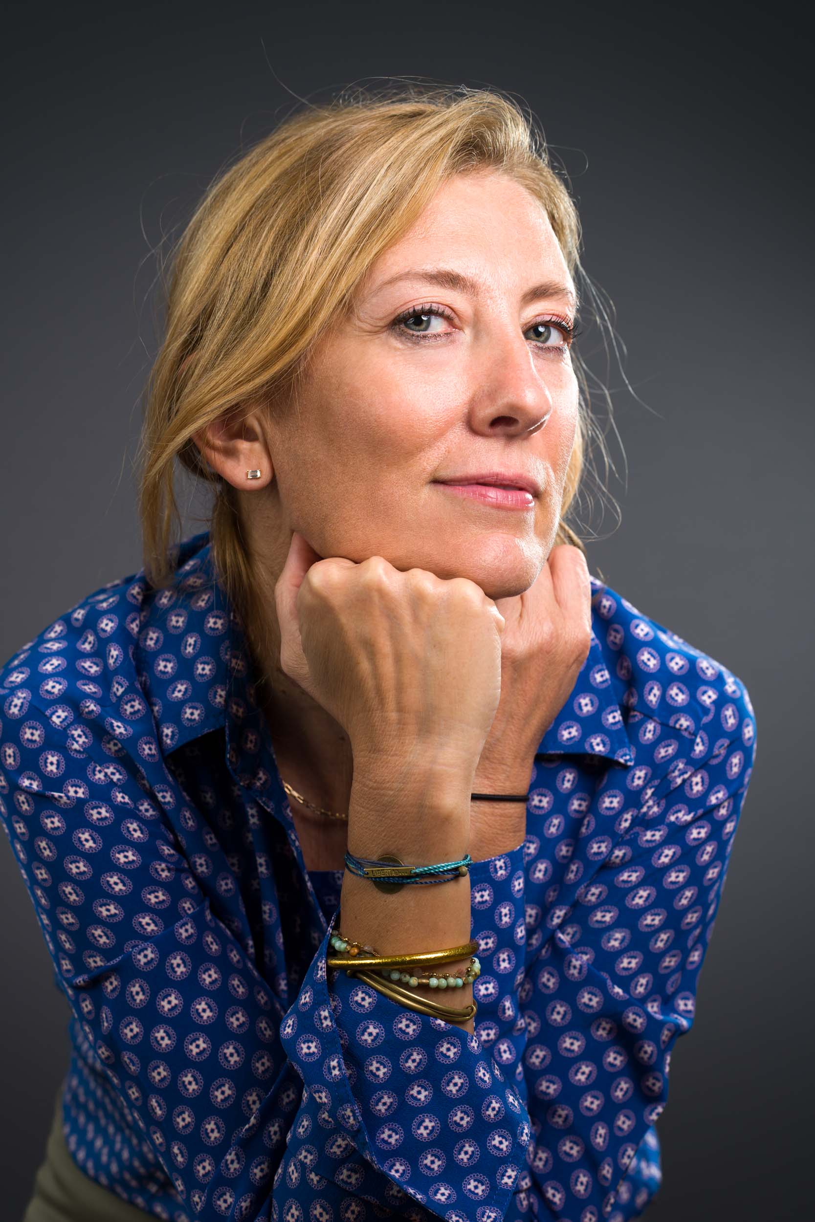 Une femme aux cheveux blonds se détache sur un fond gris foncé. Elle porte un chemisier bleu à motifs et plusieurs bracelets aux poignets. Regardant l'objectif avec assurance, le menton posé sur ses mains, elle incarne l'élégance des portraits professionnels à Bordeaux.