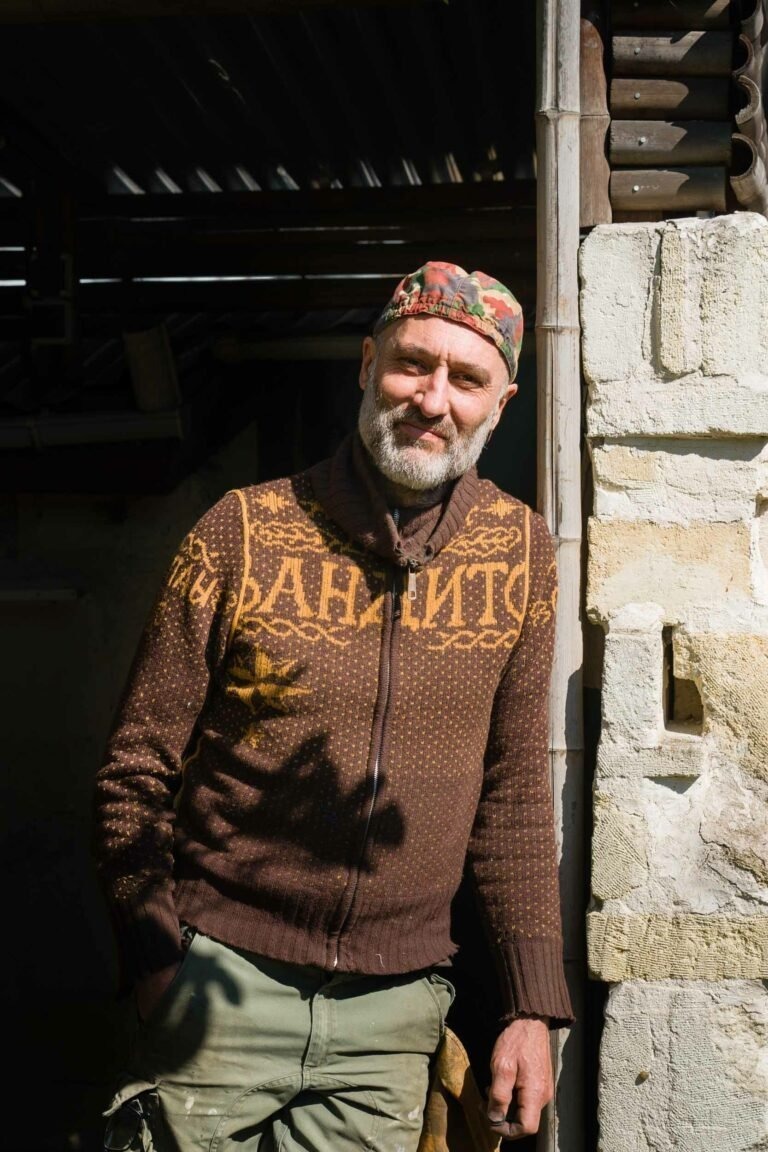 Philippe Sanchez, un coutelier à la barbe soigneusement taillée, porte un pull en maille orné de motifs complexes et une casquette camouflage. Il s'appuie contre un mur de pierre texturé, son expression détendue encadrée par la lumière du soleil qui projette des ombres sur sa tenue. L'arrière-plan révèle une structure partiellement ombragée.