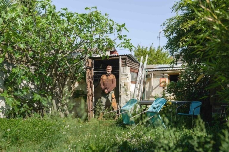 Un homme, Philippe Sanchez, se tient devant un petit cabanon en bois dans un jardin luxuriant et envahi par la végétation. La scène est remplie de verdure, notamment un figuier à gauche et diverses plantes et fleurs. Deux chaises bleues et une petite table sont au premier plan, partiellement cachées par de hautes herbes. Journée ensoleillée et lumineuse.