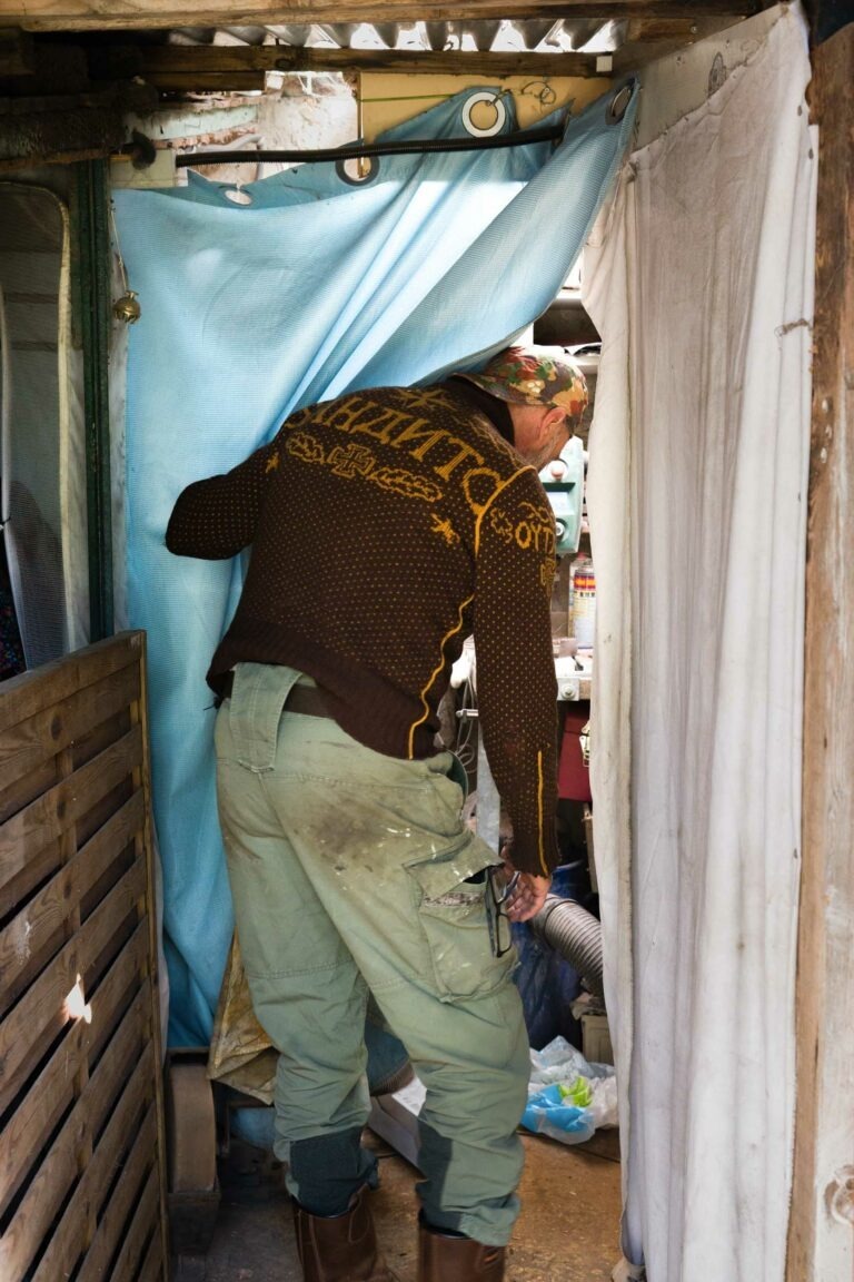 Une personne portant une veste à motifs marron et jaune, un pantalon vert et un chapeau fleuri se tient debout devant un rideau bleu. L'individu est légèrement penché, dos à la caméra, dans ce qui semble être un décor intérieur rustique et encombré rappelant le style éclectique du couturier Philippe Sanchez avec des cloisons en bois et en tissu.