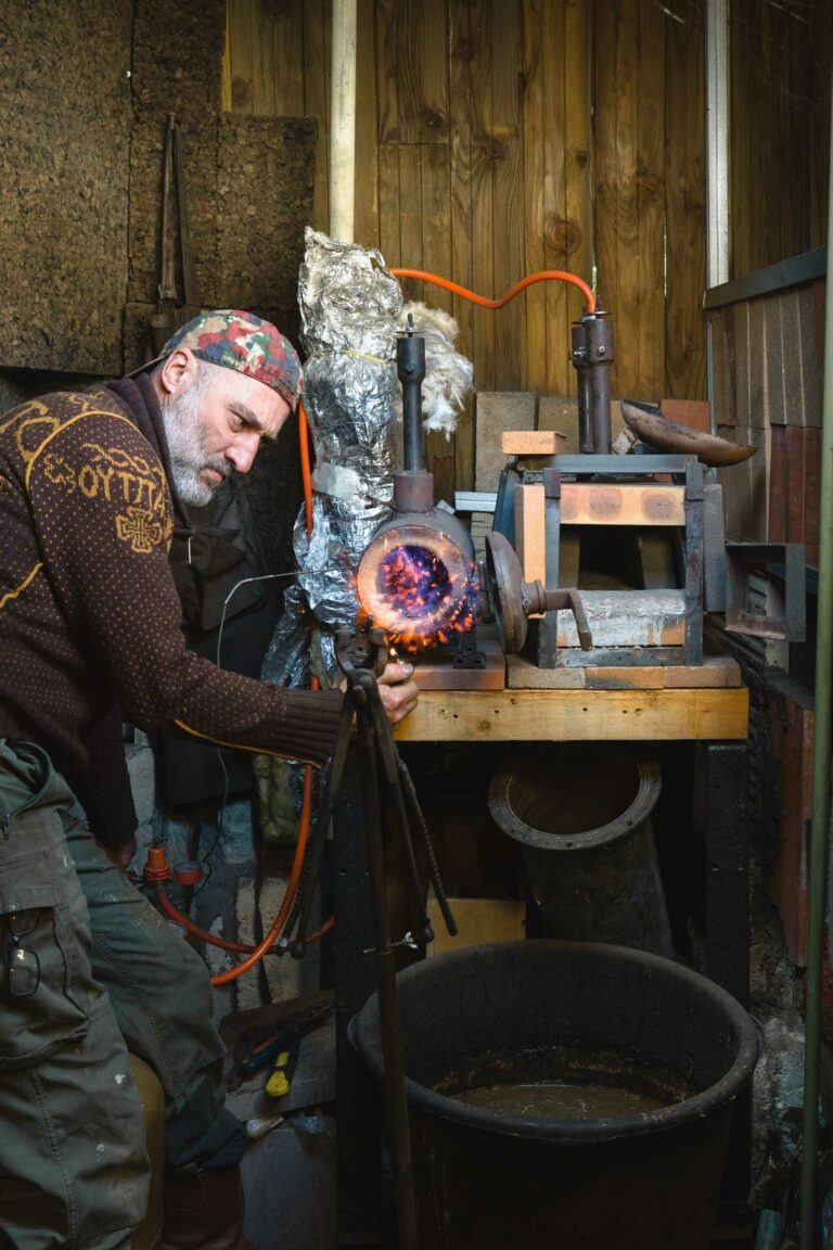 Un homme barbu, vêtu d'un pull à motifs et d'un foulard sur la tête, rappelant le style du couturier Philippe Sanchez, fait fonctionner un four dans un atelier. Il tient une boule de verre incandescente, partiellement fondue, avec des pinces, devant un établi équipé de divers outils et machines. Les murs en bois et le décor de l'atelier ajoutent une ambiance rustique au décor.