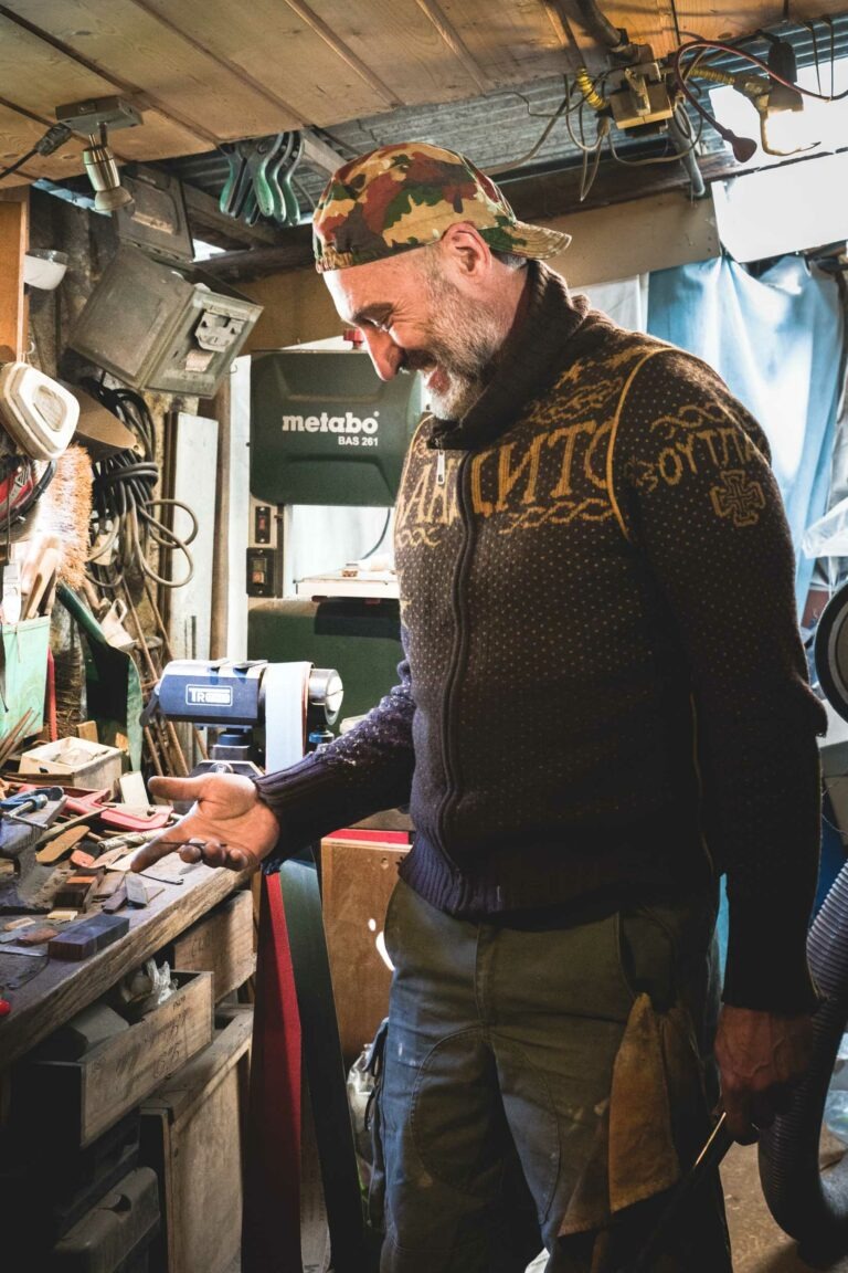 Dans l'atelier encombré du couturier Philippe Sanchez, un homme barbu, vêtu d'un pull en tricot et d'un foulard camouflage, se tient debout. Il tient un outil et examine un objet en bois posé sur un établi rempli d'outils et de matériaux divers. Des étagères avec du matériel sont visibles à l'arrière-plan, à côté d'une boîte à outils verte Metabo.