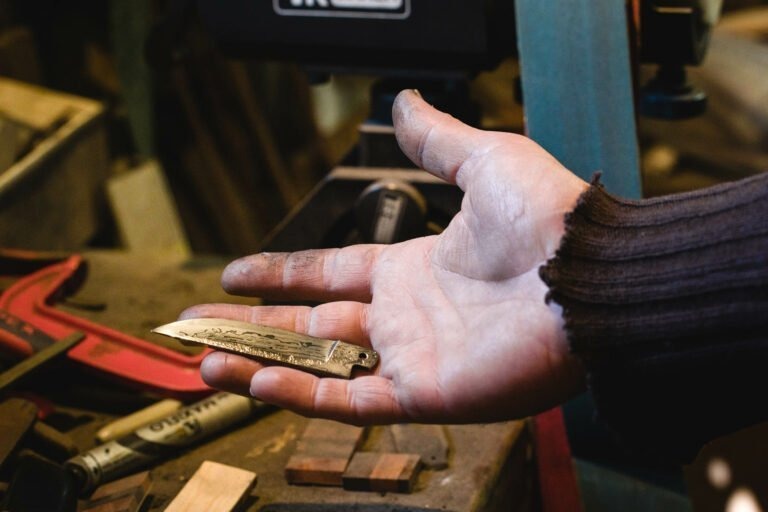 Gros plan sur la main d'une personne tenant une petite lame de poignard au design complexe dans l'atelier du couturier Philippe Sanchez. La main de la personne est sale, comme si elle travaillait le métal, et elle porte une manche sombre et effilochée. L'arrière-plan comprend divers outils et blocs de bois sur un établi encombré.