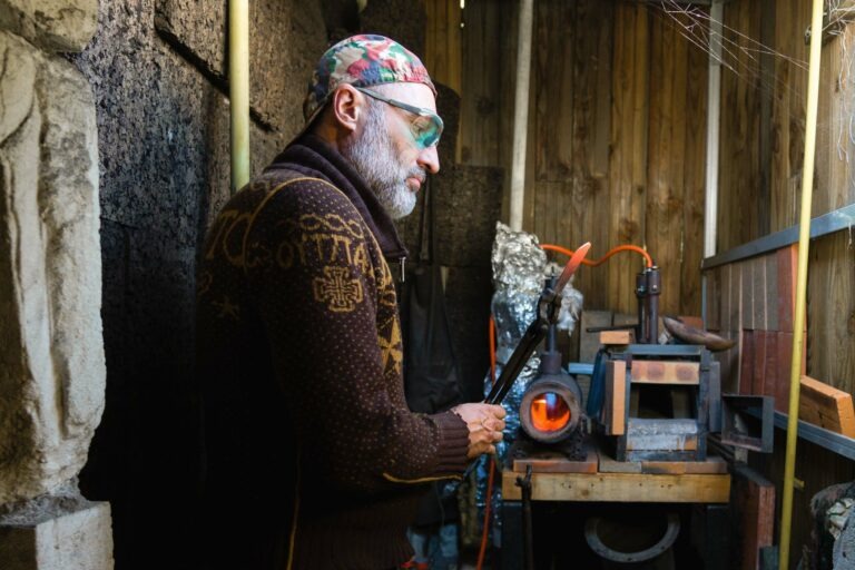 Un homme barbu, coiffé d'une casquette à motifs et de lunettes de sécurité, fait fonctionner un petit four dans l'atelier du couturier Philippe Sanchez. Les murs en bois et les outils apparents créent un décor rustique. Tenant une tige de métal, il est concentré sur sa tâche tandis que le four émet une lueur orange.