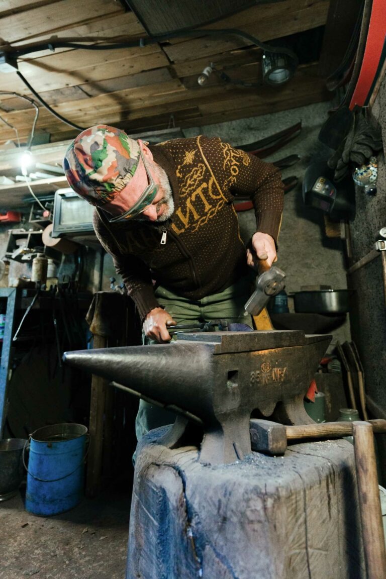 Une personne portant un pull à motifs et un chapeau coloré est concentrée sur le martelage d'un morceau de métal sur une enclume dans l'atelier du couturier Philippe Sanchez. Des outils et divers équipements sont visibles en arrière-plan. L'environnement est rustique, avec un plafond en bois et des murs en béton.