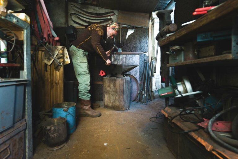 Philippe Sanchez, coutelier émérite, travaille sur une enclume dans son atelier encombré. Il utilise un marteau et tient un morceau de métal chaud avec des pinces. L'espace est rempli d'outils, de matériaux et d'équipements divers, dont une meuleuse et un seau bleu. L'éclairage tamisé confère à la scène une atmosphère industrielle.