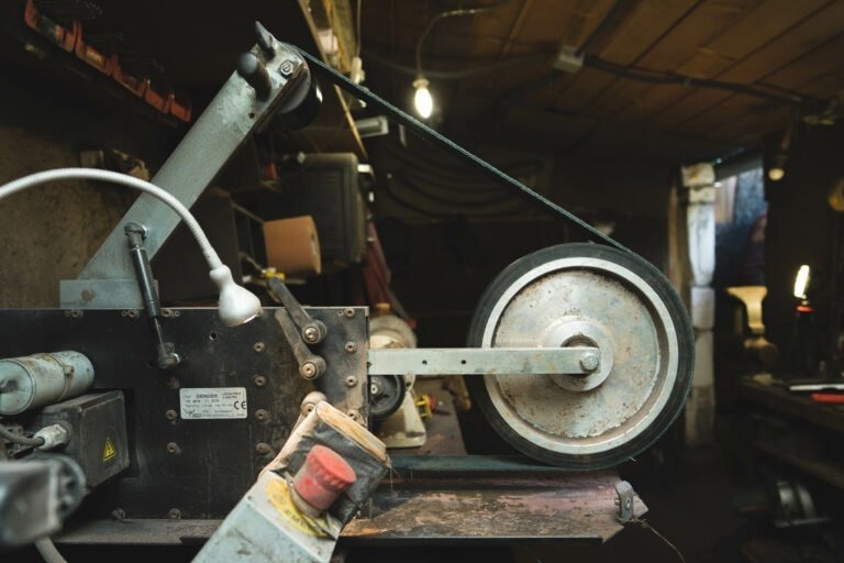 Gros plan d'une ponceuse à bande industrielle dans un atelier faiblement éclairé. La ponceuse, essentielle au métier de couturier Philippe Sanchez, se compose d'une grande roue et d'une bande, de divers boutons de réglage et d'un bouton d'arrêt d'urgence rouge. La zone environnante est encombrée d'outils et d'équipements, éclairés par une seule ampoule suspendue.