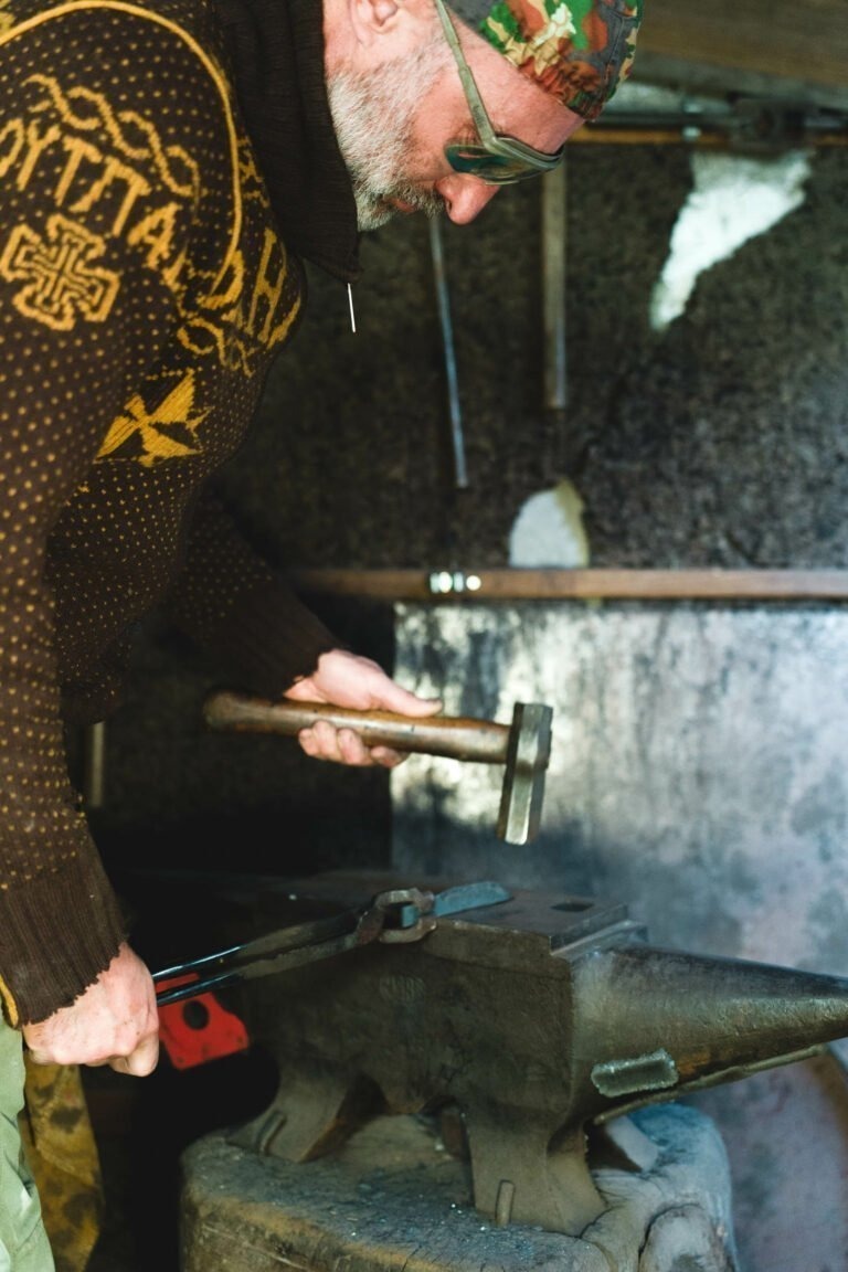 Un homme portant un pull à motifs, une casquette camouflage et des lunettes de soleil utilise un marteau et des pinces pour travailler sur un morceau de métal placé sur une enclume. L'arrière-plan montre l'atelier rustique du coutelier Philippe Sanchez, avec divers outils accrochés au mur.