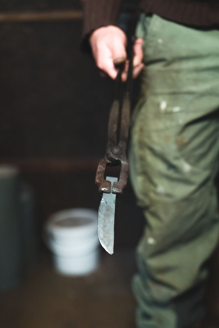 Philippe Sanchez, un coutelier expérimenté, porte un pantalon vert et une chemise à manches longues sombre, tenant un morceau de métal avec des pinces. Le métal semble être une lame ou un outil en cours de forgeage. L'arrière-plan est flou, avec un seau blanc visible sur le sol.