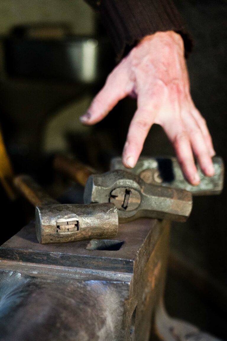 Dans l'atelier faiblement éclairé du couturier Philippe Sanchez, une main tendue vers l'un des nombreux marteaux en métal posés sur une enclume. Les outils semblent usés par l'usage et un marteau à manche en bois est visible à l'arrière-plan. La scène évoque l'art du travail du métal ou de la forge.