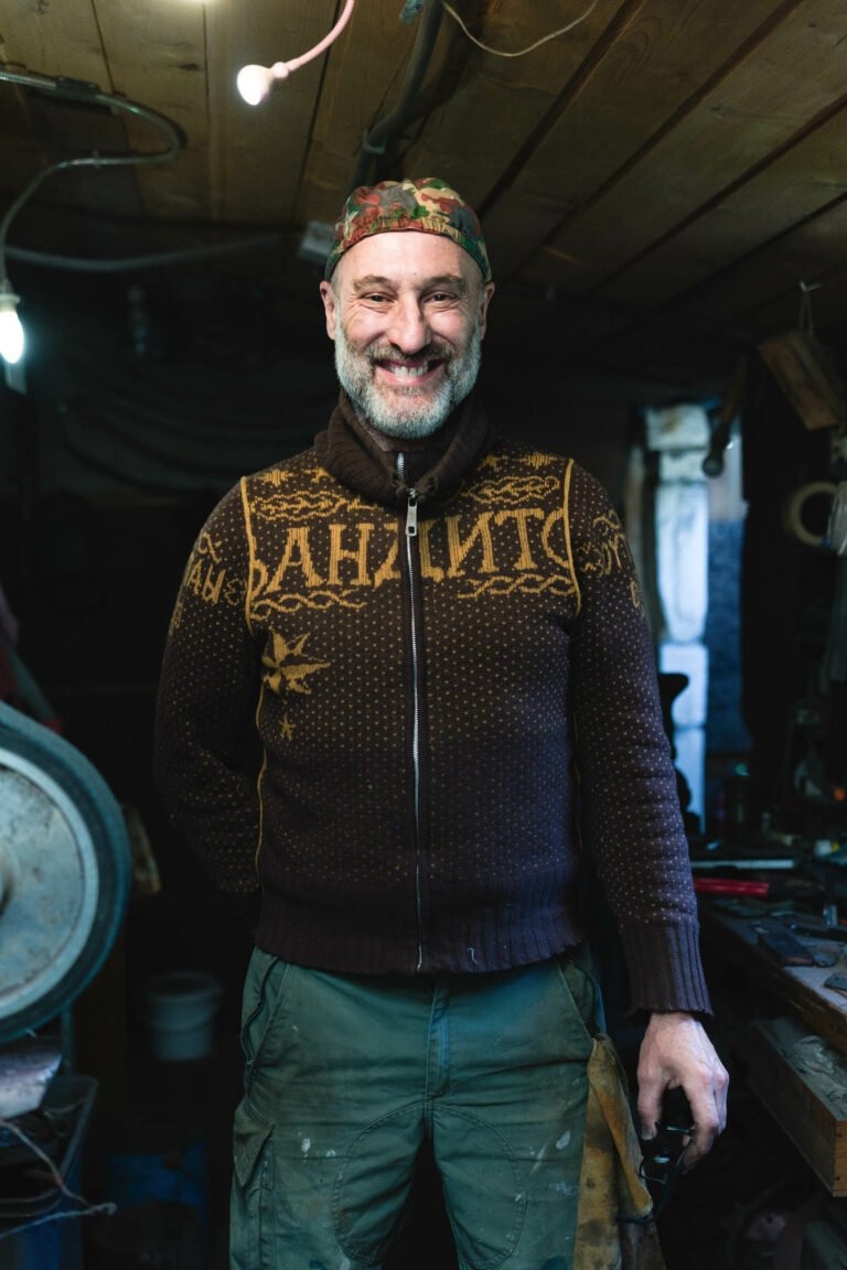 Un homme à la barbe grisonnante se tient debout, souriant, dans un atelier. Il porte un pull marron aux motifs complexes, un pantalon cargo vert et un bandeau à motifs rouges. L'atelier faiblement éclairé du couturier Philippe Sanchez présente des outils et des machines en arrière-plan, tandis qu'une seule ampoule suspendue illumine la scène.