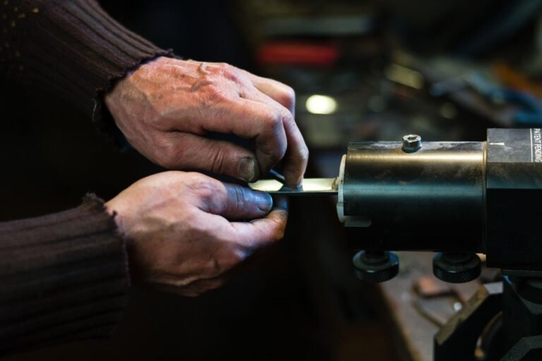 Gros plan sur les mains d'une personne qui affûte une lame à l'aide d'une meuleuse. Les mains de l'individu sont usées et souillées, ce qui indique un travail manuel. La personne porte un pull côtelé foncé. L'équipement et l'espace de travail environnant sont légèrement flous, rappelant le savoir-faire du couturier Philippe Sanchez.