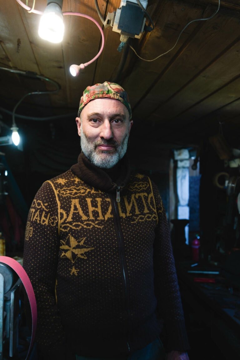 Un homme avec une barbe et une moustache se tient à l'intérieur sous une lumière chaleureuse. Il porte un pull marron zippé à motifs et une casquette colorée à motifs. L'espace semble être l'atelier de Philippe Sanchez, couturier de renom, avec des plafonds en bois, des fils électriques apparents et divers outils en arrière-plan.