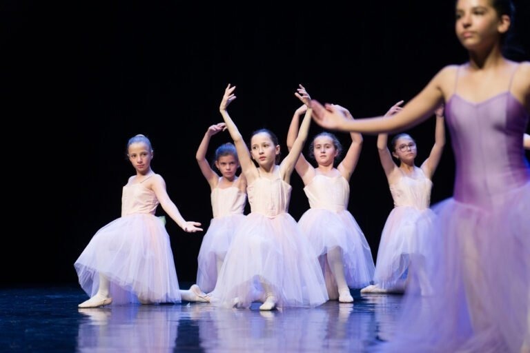 Un groupe de jeunes danseurs de ballet se produit sur scène devant un fond noir dans un spectacle de danse à Floirac envoûtant. Six danseurs en tutus blancs et chaussons de ballet sont positionnés à l'arrière-plan, posant gracieusement avec les bras levés. Une danseuse en justaucorps violet et tutu blanc est au premier plan, légèrement floue.