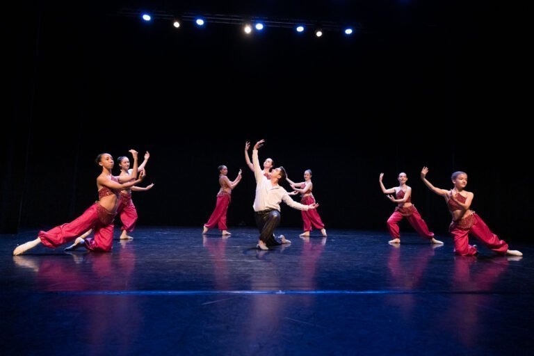 Un groupe de six danseurs se produit sur une scène faiblement éclairée, créant un spectacle de danse à Floirac. Cinq danseurs portent des costumes rouges tandis que le danseur central se distingue par son haut blanc et son pantalon noir. Ils s'agenouillent astucieusement et étendent leurs bras de manière expressive, tandis que des lumières bleues illuminent la scène. Le sol de la scène reflète les lumières.