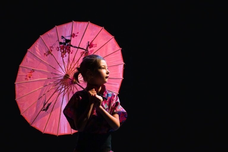 Une jeune fille en tenue traditionnelle tient debout un grand parasol rouge orné de motifs de fleurs de cerisier. Elle regarde sur le côté et un projecteur du Spectacle de danse à Floirac l'illumine sur un fond sombre, mettant en valeur les couleurs vives et les détails complexes du parasol.