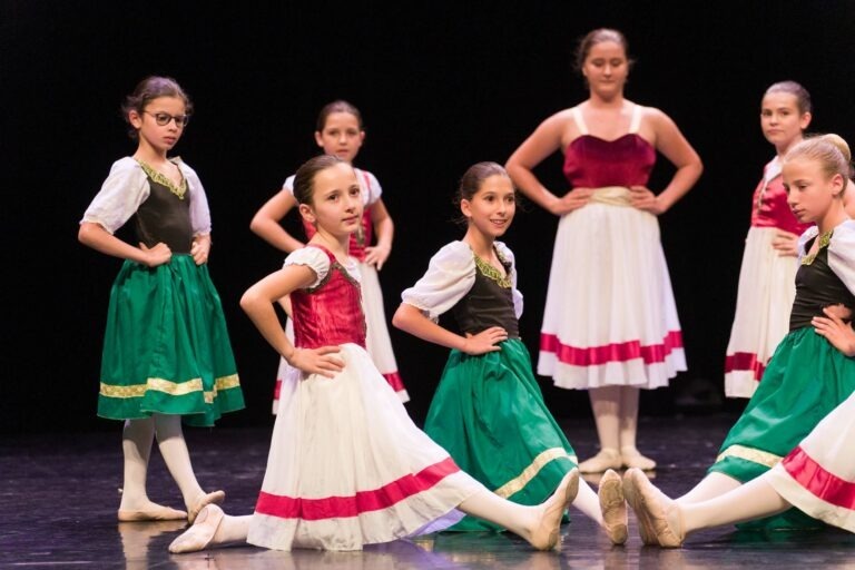 Un groupe de jeunes danseurs de ballet en costumes traditionnels se produit sur scène, vêtus de robes blanches avec des jupes vertes et des corsages marron. Certains danseurs se tiennent debout, les mains sur les hanches, tandis que d'autres sont à genoux, une jambe tendue. La scène, préparée pour le spectacle de danse à Floirac, est faiblement éclairée par un fond sombre.