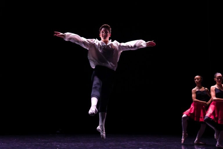 Un danseur de ballet en chemise blanche et pantalon noir exécute un saut, les bras tendus, au centre de la scène, sur un fond sombre. Deux danseuses en jupe rouge et haut noir se tiennent à l’arrière-plan sur le côté droit, partiellement éclairées. C’est vraiment un spectacle de danse à Floirac fascinant.