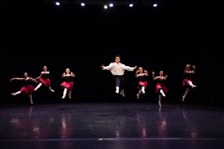 Six danseurs de ballet sautent sur scène devant un fond sombre. Cinq danseuses portent des hauts blancs et des jupes rouges, tandis qu'un danseur, au centre, porte une chemise blanche et un pantalon noir. Cette scène fascinante du Spectacle de danse à Floirac est illuminée par une rangée de lumières au-dessus d'eux.