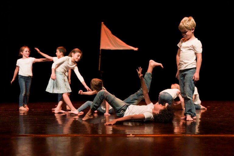 Lors du spectacle de danse à Floirac, un groupe d'enfants se produit sur une scène sombre avec un tissu blanc en forme de triangle en arrière-plan. Certains enfants sont debout, se tendent les mains et se tiennent, tandis que d'autres posent au sol. Ils sont pieds nus et vêtus de hauts blancs décontractés et de jeans, créant une scène vivante et expressive.