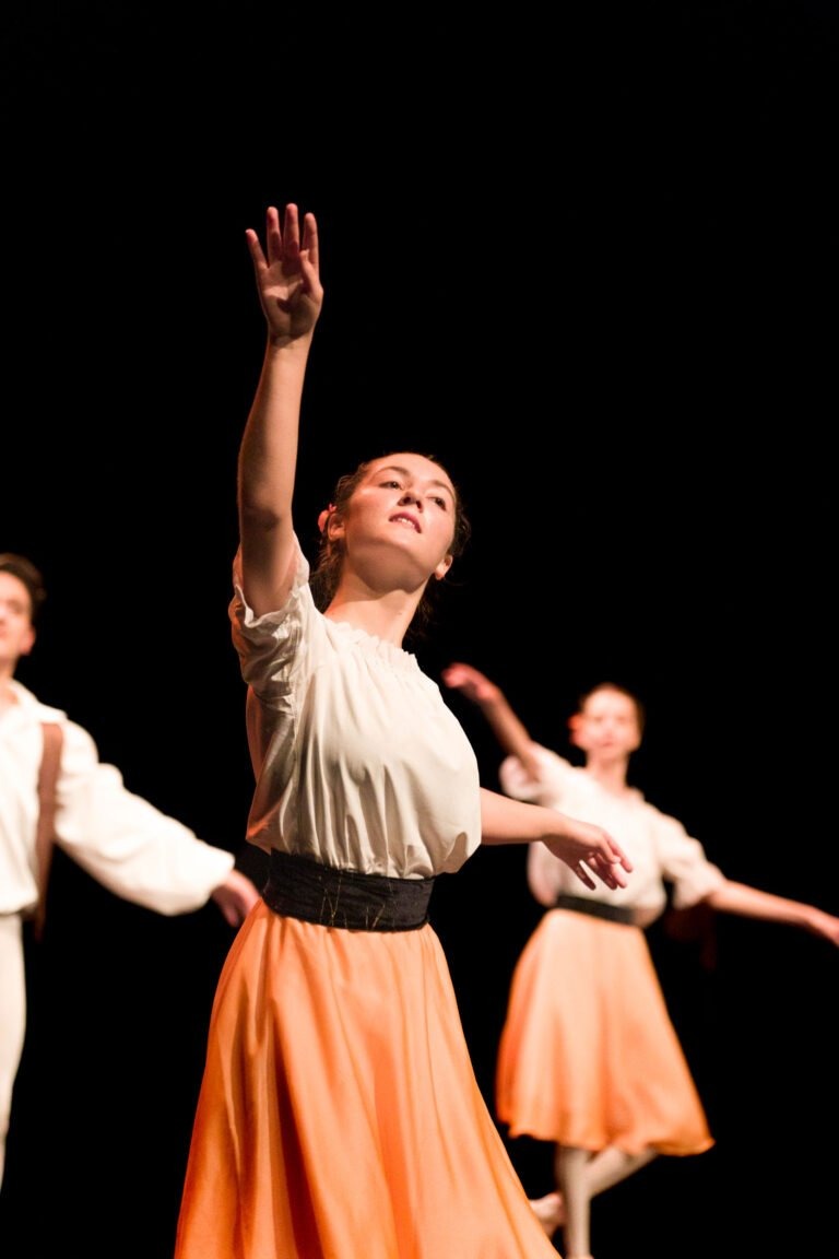 Une ballerine vêtue d'une blouse blanche et d'une jupe orange se produit sur scène, le bras tendu vers le haut, lors du Spectacle de danse à Floirac. Une autre danseuse, habillée de la même manière, est visible à l'arrière-plan. Les danseuses sont éclairées sur un fond sombre, ce qui met en valeur leurs mouvements gracieux.