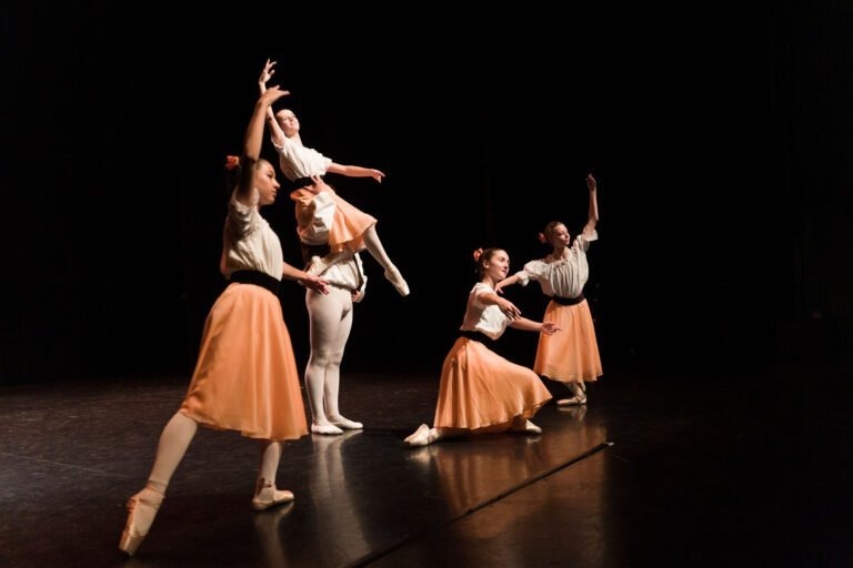 Quatre danseurs de ballet se produisent sur scène sur fond sombre dans le spectacle de danse à Floirac. Trois femmes en hauts beiges et jupes pêche fluides et un homme en tenue blanche sont photographiés dans des poses élégantes. Un danseur est soulevé par l'homme, tandis que les autres prennent des positions équilibrées, mettant l'accent sur la grâce et l'équilibre.