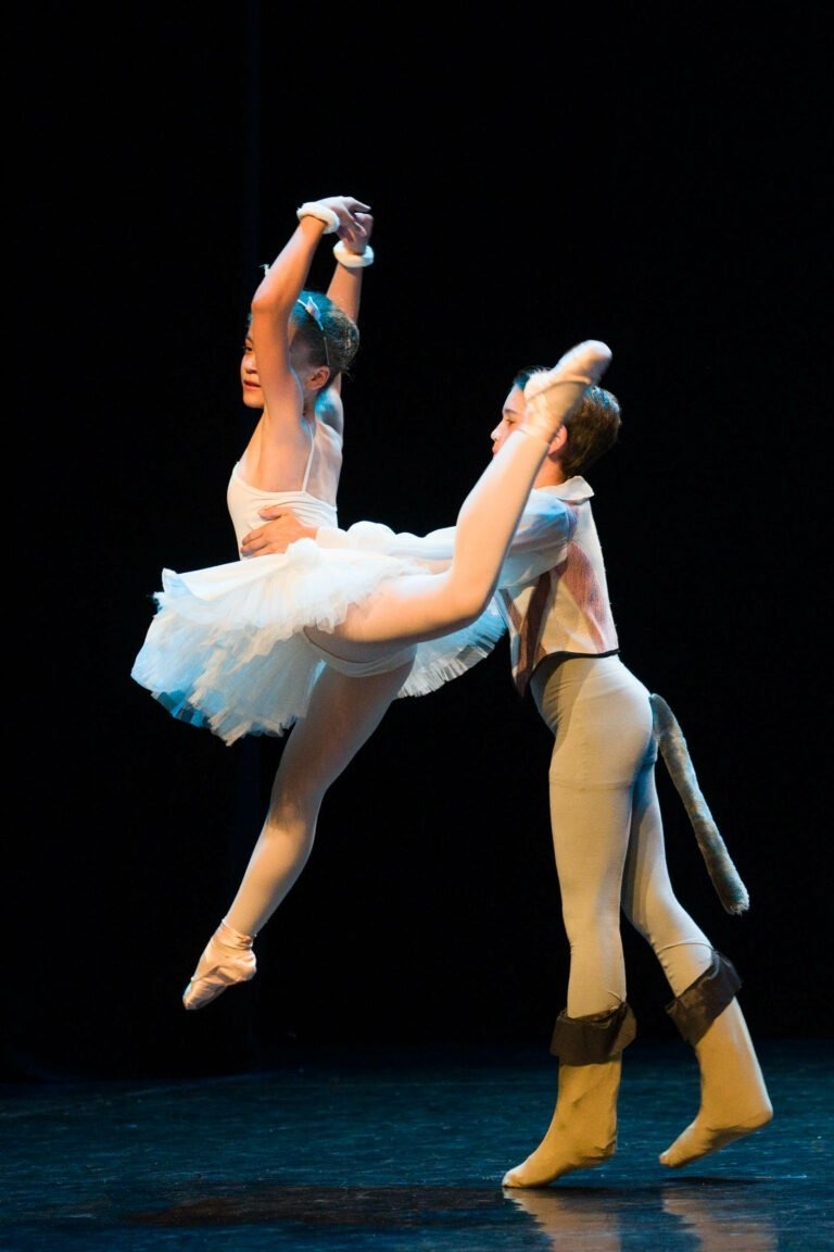 Deux jeunes danseurs de ballet se produisent sur scène sur un fond sombre. Le danseur de droite, vêtu d'un costume de souris, soulève dans les airs le danseur de gauche, qui porte un tutu blanc et des chaussons de ballet. Les deux danseurs ont des expressions concentrées pendant ce spectacle de danse à Floirac envoûtant.