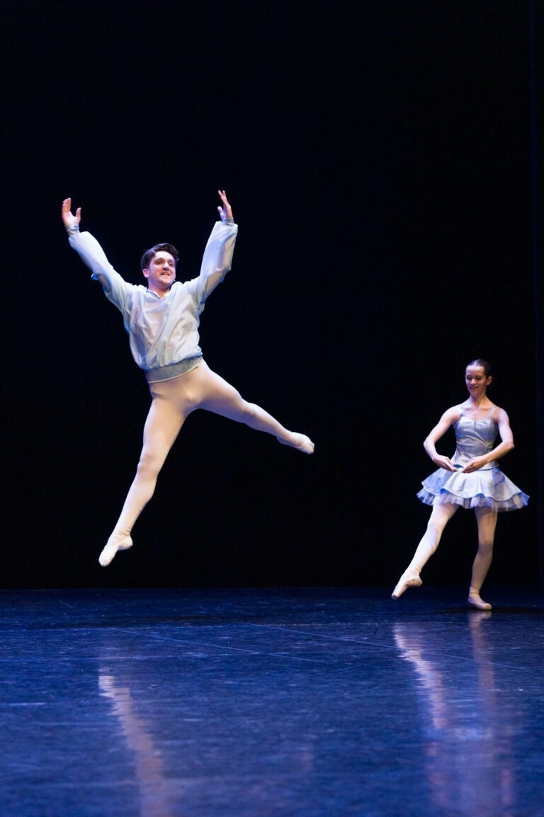 Un danseur de ballet en tenue blanche saute gracieusement dans les airs sur une scène sombre, les bras écartés. Une danseuse en tutu bleu clair et chaussons de pointe se tient debout, le pied droit en pointe, la jambe gauche légèrement pliée. Tous deux semblent concentrés et élégants, incarnant l'essence du Spectacle de danse à Floirac.