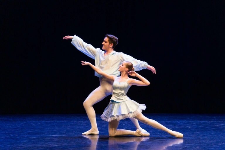 Un danseur et une danseuse se produisent sur une scène faiblement éclairée au Spectacle de danse à Floirac. Le danseur, vêtu d'une chemise blanche ample et de collants, se tient debout en position de fente avec un bras tendu. La danseuse, vêtue d'un tutu blanc court, est à genoux, l'autre jambe tendue derrière elle et un bras levé.