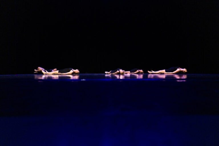 Quatre danseurs en tenue noire sont allongés sur le dos, en ligne horizontale, sur une scène sombre où se reflète une lumière bleu foncé. La scène minimaliste du Spectacle de danse à Floirac met en valeur la symétrie et l'immobilité des positions des danseurs.