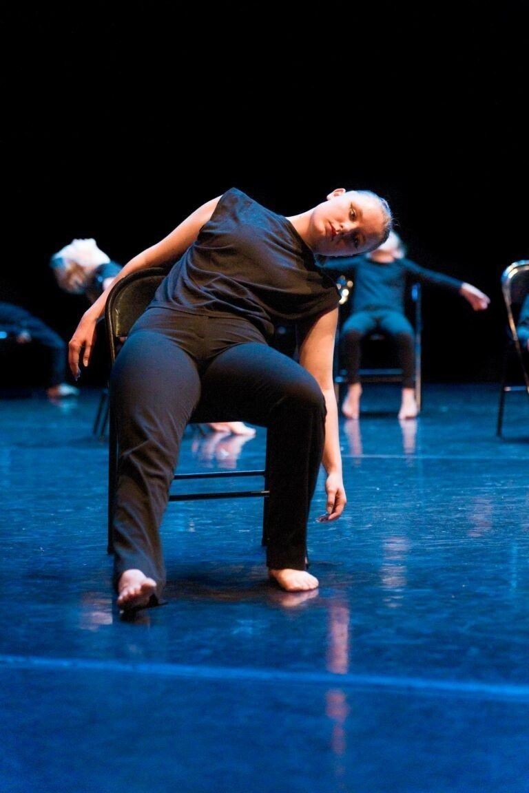 Une danseuse en tenue noire est assise sur une chaise sur une scène faiblement éclairée, penchée sur le côté, un bras reposant sur le dossier. D'autres danseuses sont visibles sur des chaises similaires en arrière-plan, créant un sentiment de cohésion. Le sol reflète les lumières de la scène, ajoutant de la profondeur à ce spectacle de danse à Floirac.