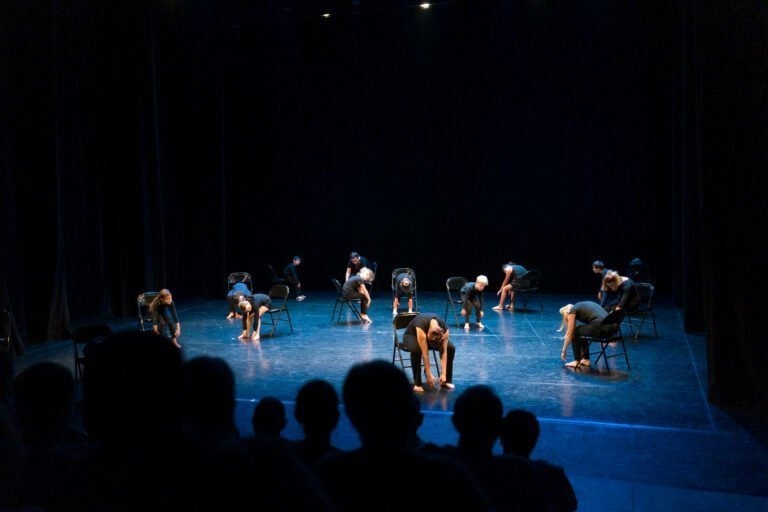 Un groupe de danseurs vêtus de noir exécute une chorégraphie sur la scène du Spectacle de danse à Floirac, chacun interagissant avec des chaises pliantes noires en guise d'accessoires. Les danseurs sont répartis sur la scène, se penchant et se tendant à l'unisson. La scène est faiblement éclairée et un public regarde au premier plan.