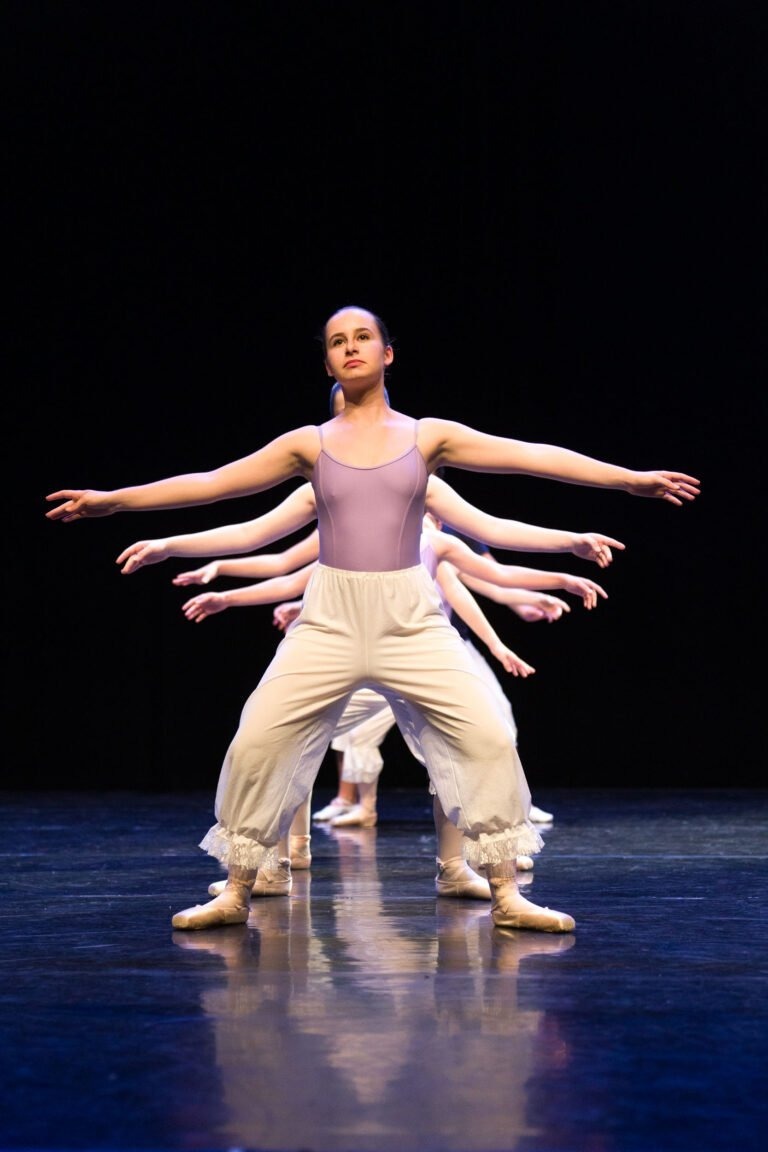 Un groupe de danseurs de ballet se produit sur scène, tous vêtus de costumes clairs, les femmes portant des justaucorps lilas et des pantalons blancs flottants. Ils sont synchronisés, les bras tendus sur les côtés et les jambes légèrement pliées. Dans ce Spectacle de danse à Floirac, le fond sombre concentre l'attention sur les danseurs.