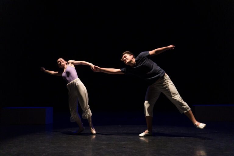 Deux danseurs se produisent sur scène sous un éclairage de projecteur dans un décor sombre au Spectacle de danse à Floirac. Tous deux portent des tenues de danse décontractées. Ils se tiennent par la main et s'écartent l'un de l'autre, créant un mouvement dynamique et fluide. L'arrière-plan est complètement sombre, ce qui concentre l'attention sur les mouvements des danseurs.