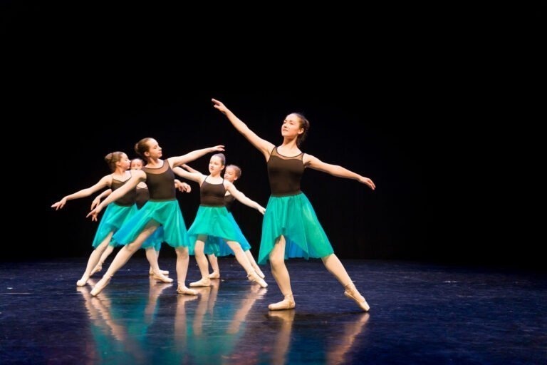 Un groupe de six jeunes ballerines se produit sur une scène au fond sombre lors du Spectacle de danse à Floirac. Elles portent des costumes assortis, composés de hauts noirs et de jupes turquoise. Toutes sont dans des poses de danse synchronisées, la plupart étendant gracieusement un bras et une jambe. Le sol de la scène reflète leurs mouvements.