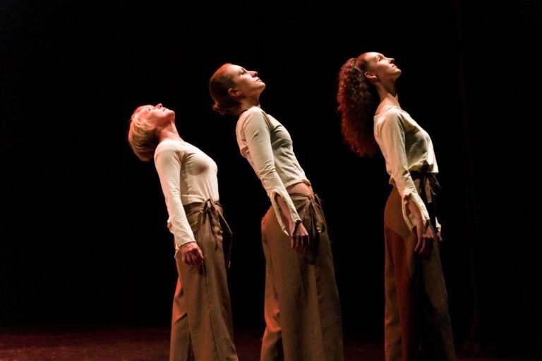 Trois femmes se tiennent en ligne sur une scène sombre du Spectacle de danse à Floirac, penchées en arrière et la tête relevée. Elles portent des hauts à manches longues couleur crème et des pantalons amples marron. L'éclairage met en valeur leurs profils sur le fond noir, créant un effet dramatique.