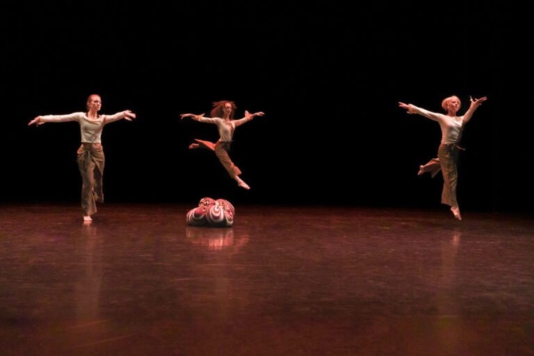 Trois danseurs en tenue décontractée, en plein saut, sur une scène sombre. Ils sont pieds nus et chacun prend une pose différente, les bras et les jambes étendus. En dessous d'eux, un objet coloré et tourbillonnant repose sur le sol poli, captant la lumière. Ce moment spectaculaire fait partie du Spectacle de danse à Floirac, avec un fond noir qui met en valeur les mouvements des danseurs.