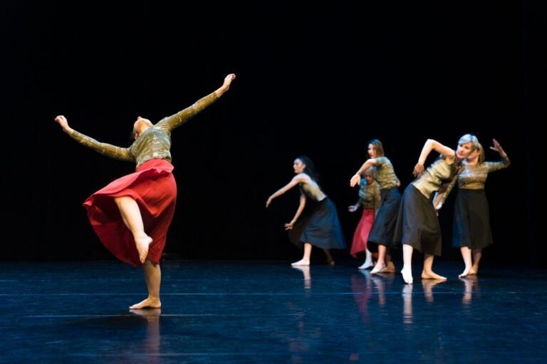 Au Spectacle de danse à Floirac, cinq danseurs se produisent sur scène, vêtus de hauts à manches longues et de jupes de différentes couleurs sombres, dont une rouge. Le danseur au premier plan lève haut sa jambe gauche et se cambre vers l'arrière tandis que les autres adoptent des poses dynamiques et expressives. La scène est sombre, ce qui met en valeur leurs mouvements.