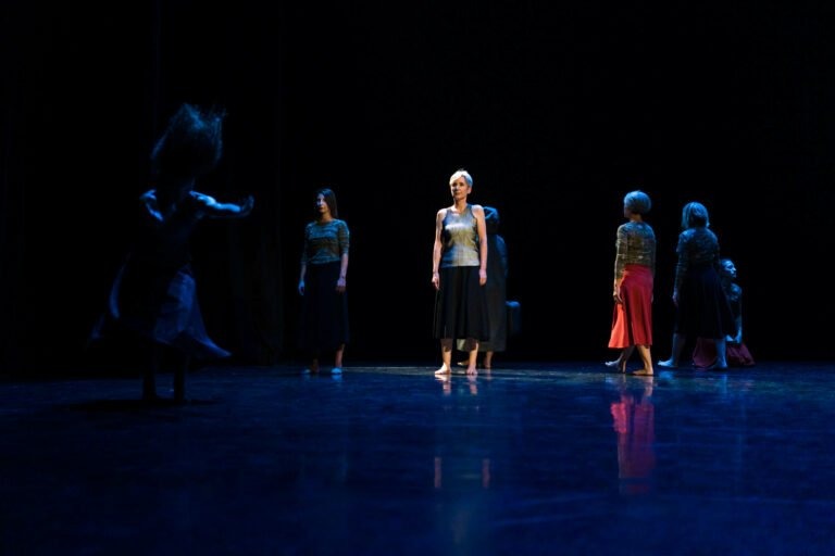 Un groupe de danseurs exécute une danse contemporaine sur une scène sombre du Spectacle de danse à Floirac. Un danseur en blanc et noir est bien éclairé, debout au centre. D'autres danseurs vêtus de divers costumes sombres sont dispersés autour, certains tournés vers l'extérieur. Un danseur à gauche semble être en mouvement, avec une teinte bleue mettant en valeur la scène.