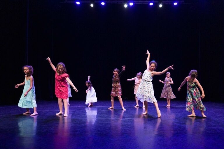 Un groupe de huit enfants exécute une danse sur une scène faiblement éclairée. Les danseurs portent des robes colorées et sont pieds nus. Des lumières bleues les éclairent d'en haut, soulignant leurs poses variées et leurs expressions de grâce et de joie. Le fond est sombre, mettant en valeur les danseurs de ce Spectacle de danse à Floirac.