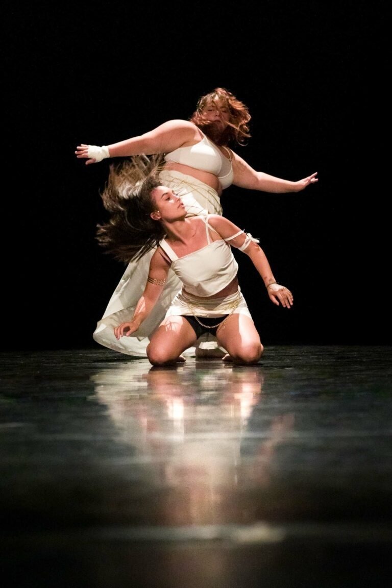 Dans un spectacle de danse à Floirac, deux danseurs se produisent sur scène devant un fond noir. L'un est à genoux, la tête penchée en arrière et les cheveux flottants, tandis que l'autre se tient debout derrière, penché en arrière avec grâce, les bras écartés. Tous deux portent des costumes blancs et fluides qui reflètent la lumière. Leurs mouvements sont dynamiques et expressifs.