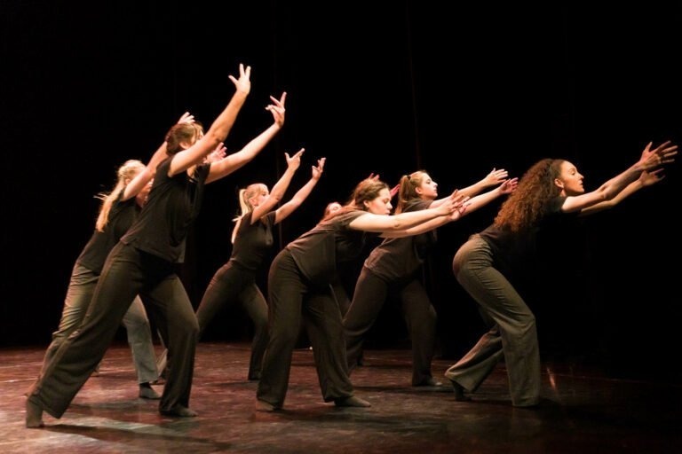 Six danseurs en tenue sombre se produisent sur une scène faiblement éclairée au Spectacle de danse à Floirac. Ils adoptent une posture dynamique, se jetant en avant, les bras tendus vers l'extérieur et les doigts écartés, exprimant un mouvement fluide et intense. Le fond est noir, ce qui met en valeur leurs mouvements et leurs expressions.