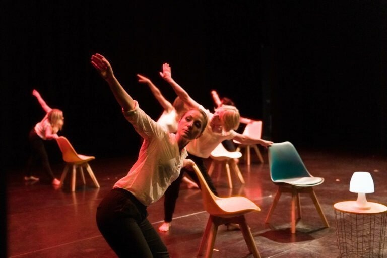 Un groupe de danseurs évolue sur une scène tamisée, illuminée par des lumières chaudes rouge et jaune. Vêtus de chemises blanches et de pantalons noirs, ils interagissent avec des chaises de style moderne et une petite table sur laquelle est posée une lampe blanche. Leurs mouvements dynamiques et expressifs captivent le public du Spectacle de danse à Floirac.