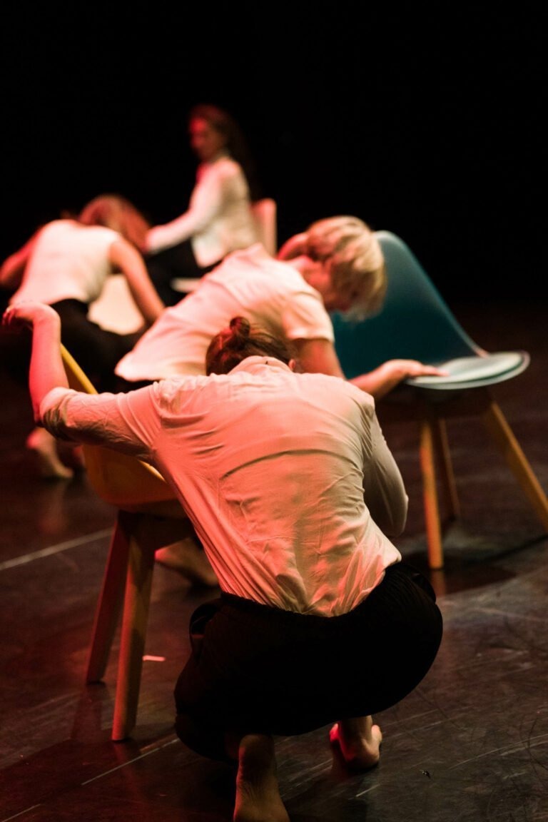 Dans le spectacle de danse à Floirac, à la lumière tamisée, quatre danseurs en chemise blanche et pantalon sombre se produisent avec des chaises. Un danseur se penche sur une chaise bleue tandis qu'un autre s'agenouille devant une chaise jaune. Deux danseurs à l'arrière-plan interagissent avec leurs chaises, ajoutant de la profondeur à la scène.