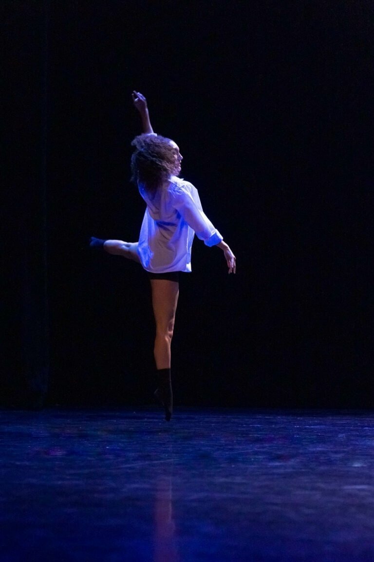 Une danseuse vêtue d'une chemise blanche et d'un short noir exécute un mouvement de ballet sur scène, en équilibre sur une jambe, l'autre tendue derrière elle. Son bras gauche est levé gracieusement au-dessus de sa tête. Le fond est sombre, mettant en valeur la silhouette élégante de la danseuse mise en lumière au Spectacle de danse à Floirac.