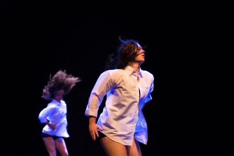 Deux danseurs se produisent sur une scène sombre. Le danseur au premier plan porte une chemise blanche et un pantalon noir, les cheveux flottant sur le côté. Un autre danseur à l'arrière-plan, également en chemise blanche, a les cheveux flottant vers l'arrière de manière dynamique. Le spectacle de danse à Floirac concentre toute l'attention sur les danseurs.