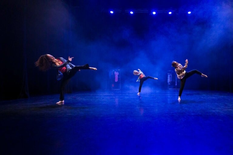 Trois danseurs évoluent sur une scène sombre illuminée de lumières bleues. Les danseurs, vêtus de costumes colorés, sont en plein mouvement avec une jambe tendue. De la brume ou de la fumée crée une atmosphère onirique sur le sol brillant, ajoutant à l'effet visuel de leur routine de danse expressive au spectacle de danse à Floirac.